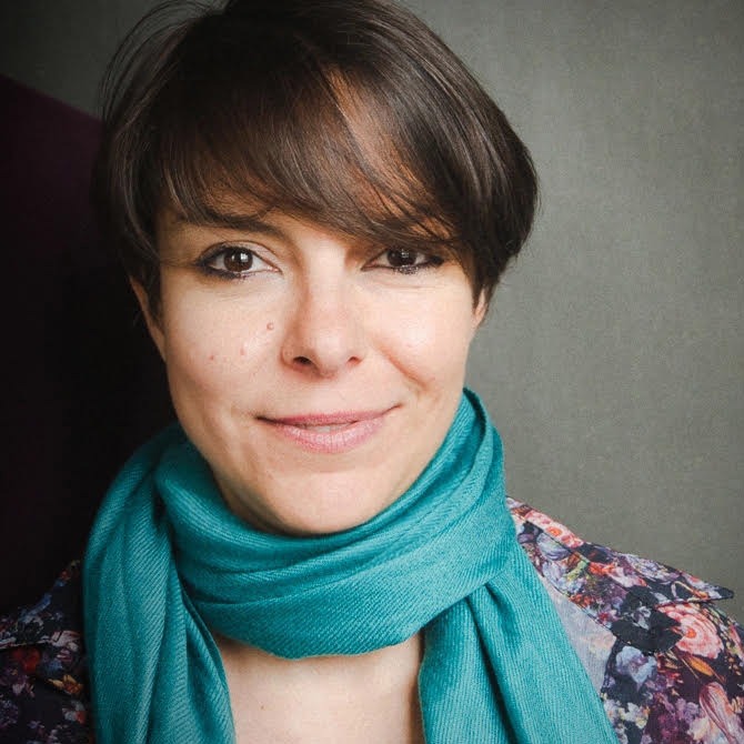 Portrait of Dalila D'Amico, smiling, with a short brown bob and a teal scarf over a floral jacket, against a grey background.