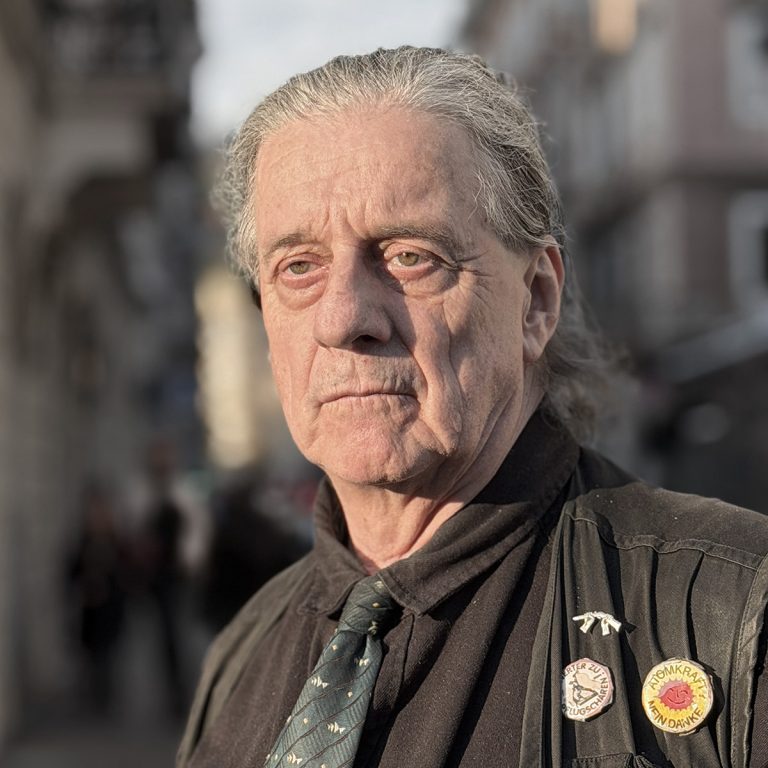 Portrait of Herbert Gantschacher on a sunlit street, wearing a dark jacket with anti-nuclear and peace badges.