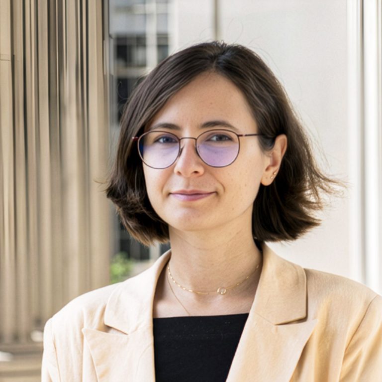 Portrait of Iris Popescu, with a dark brown bob and round glasses, wearing a cream blazer over a black top, standing among pale stone columns.