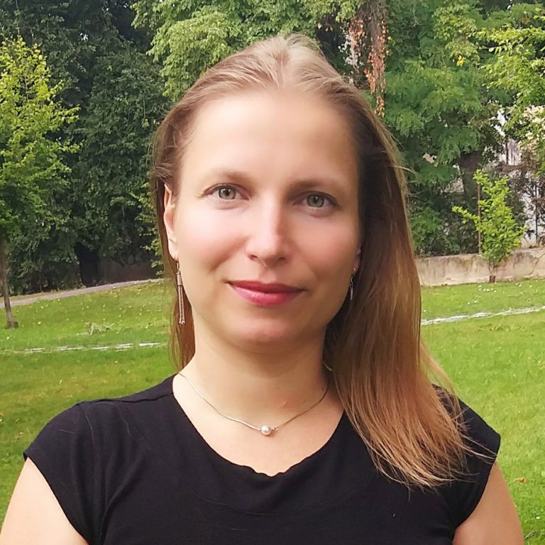 Portrait of Jitka Vrbková, smiling, with long fair hair, wearing a black top, a fine necklace and drop earrings, photographed outdoors in a green park.