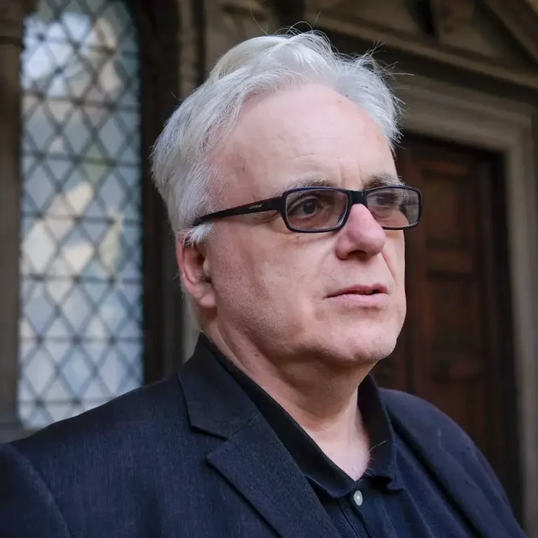 Portrait of Jonathan METH, man with white hair and black-framed glasses in a dark jacket, in front of a historic building.