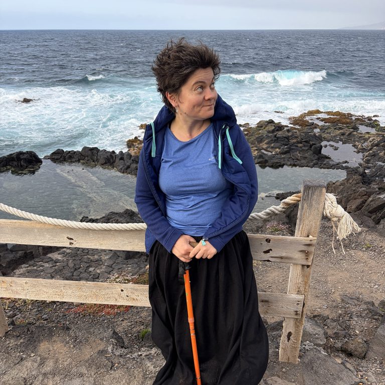 Portrait of Olga Macrinici, woman in a blue jacket and long black skirt, holding an orange walking stick, standing by a wooden fence on a rocky coastline with waves behind her.